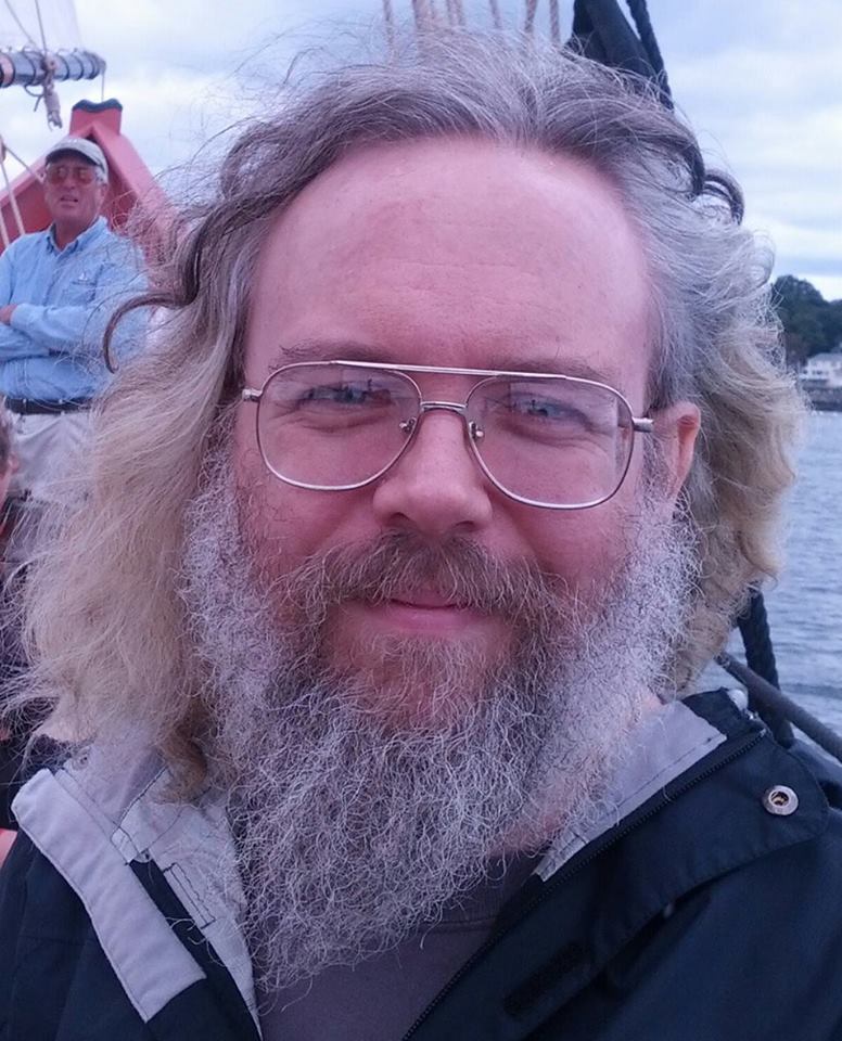 A picture of a man with long, gray hair and beard blowing in the breeze. He has large glasses and he is smiling.