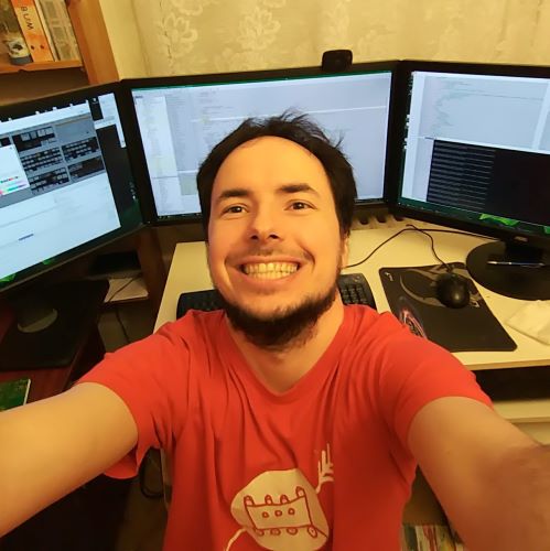 A young man with light facial hair takes a picture of himself with three computer monitors behind him. He has a huge smile on his face