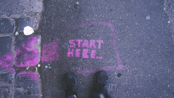 Looking down, you can see someone's feet, on concrete next to a purple spraypainted lettering that says 'Start Here'