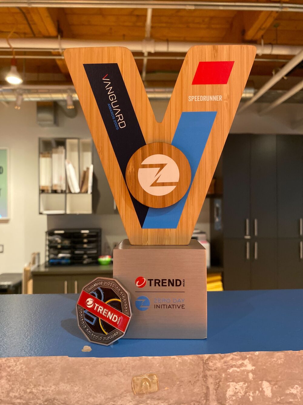 Photo of the trophy. It looks like a big V-letter shaped wooden block, on top of a metal foot. The left side of the V says Vanguards Awards 2025 and the right side says Speedrunner. There is also text indicating this was awarded by the Trend Micro Zero Day Initiative (ZDI)
