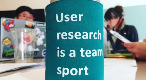 A soda can in a coozy that says "User research is a team sport," sitting on a table with people & laptops in the background.