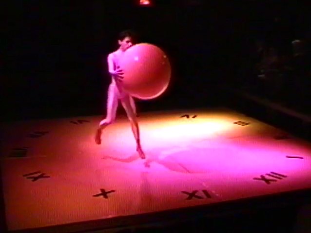 A man in a white leotard holding a large yoga ball on a small stage.