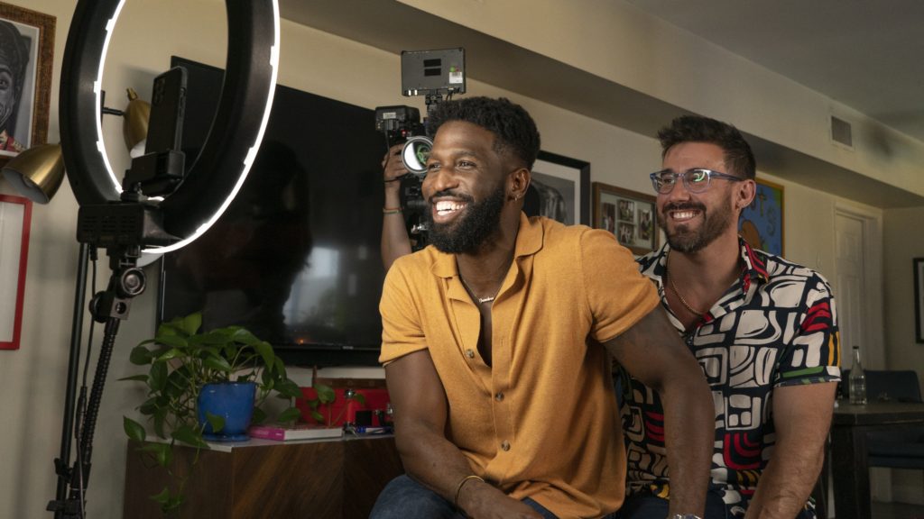 Brandon Kyle Goodman and their husband Matthew sit next to each other, smiling in front of a phone and a ring light.