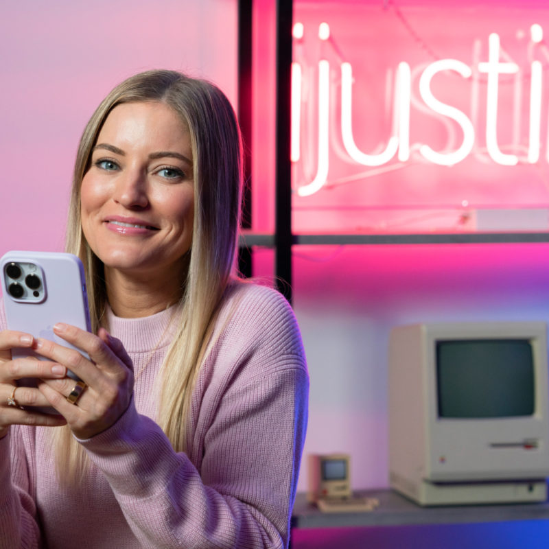 Justine Ezarik holds her phone as she smiles at the camera. In the background, a neon sign reads ijustine.