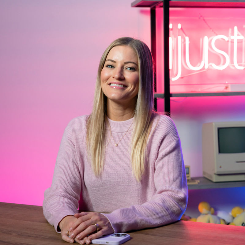 Justine Ezarik smiles at the camera. In the background, a neon sign reads ijustine.
