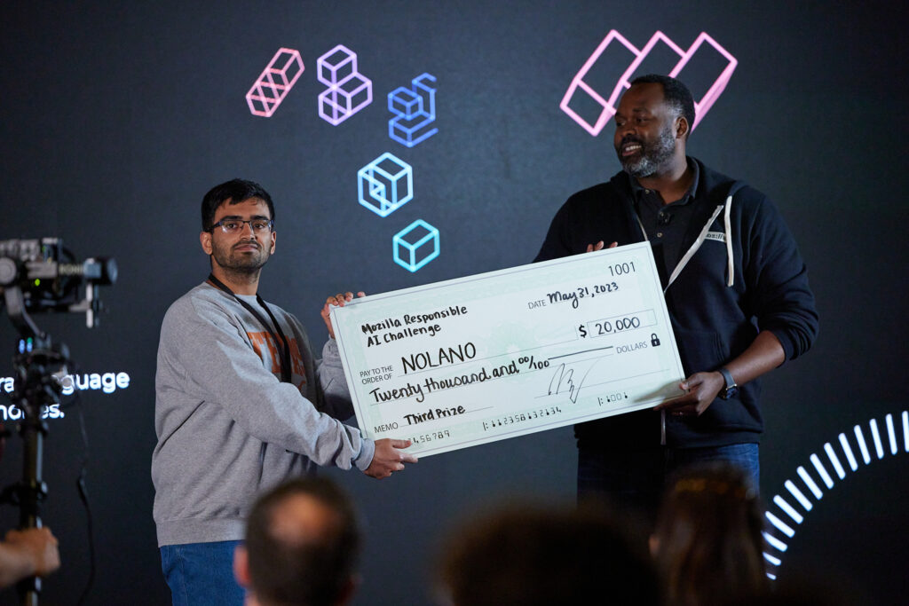 Nolano co-founder Ayush Kaushal and Imo Udom, Mozilla's SVP of innovation ecosystems, hold a check on the Mozilla AI Challenge event stage.