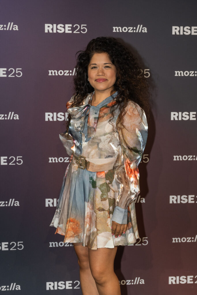 A woman with long, curly hair stands in front of a backdrop featuring the logos "RISE25" and "mozilla". She is wearing a colorful, patterned dress with a pleated skirt and puffed sleeves. The dress has a belt at the waist, and she is smiling slightly at the camera.