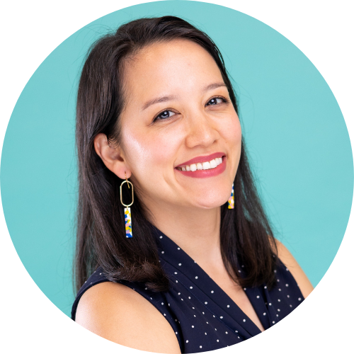 A smiling woman with long dark hair, wearing colorful earrings and a navy blue polka dot top, in front of a turquoise background.