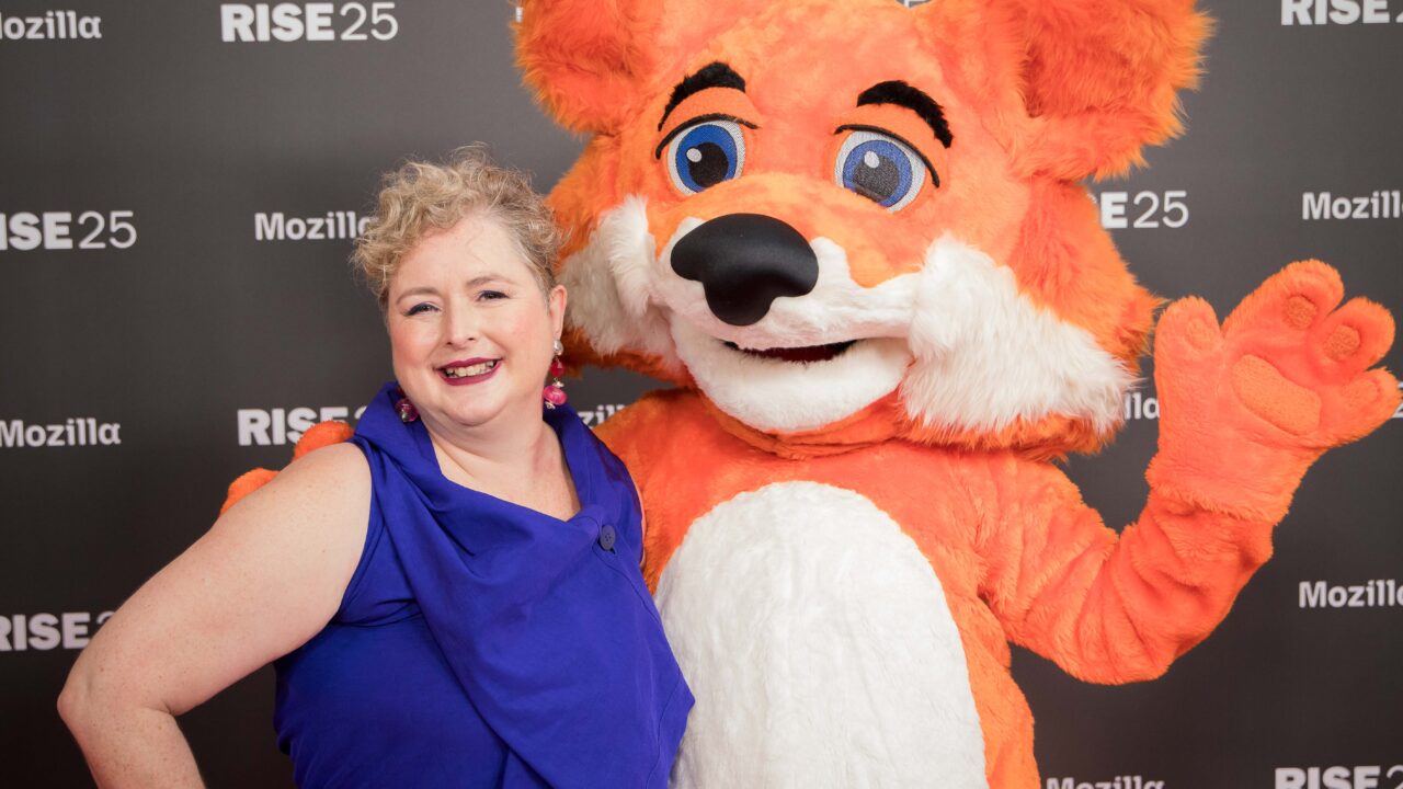 A smiling woman in a blue dress poses with the Firefox mascot at a Mozilla event with a "RISE25" backdrop.