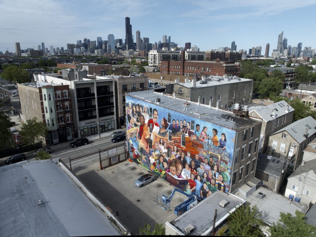 A colorful mural on a building in Chicago's Pilsen neighborhood, featuring diverse faces and scenes from the community. The Chicago skyline looms in the background under a bright, clear sky.