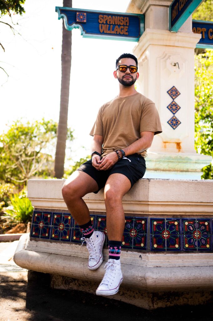 A man with sunglasses and a beard sits casually on a stone bench in front of a sign that reads "Spanish Village." He is wearing a beige t-shirt, black shorts, and white sneakers.