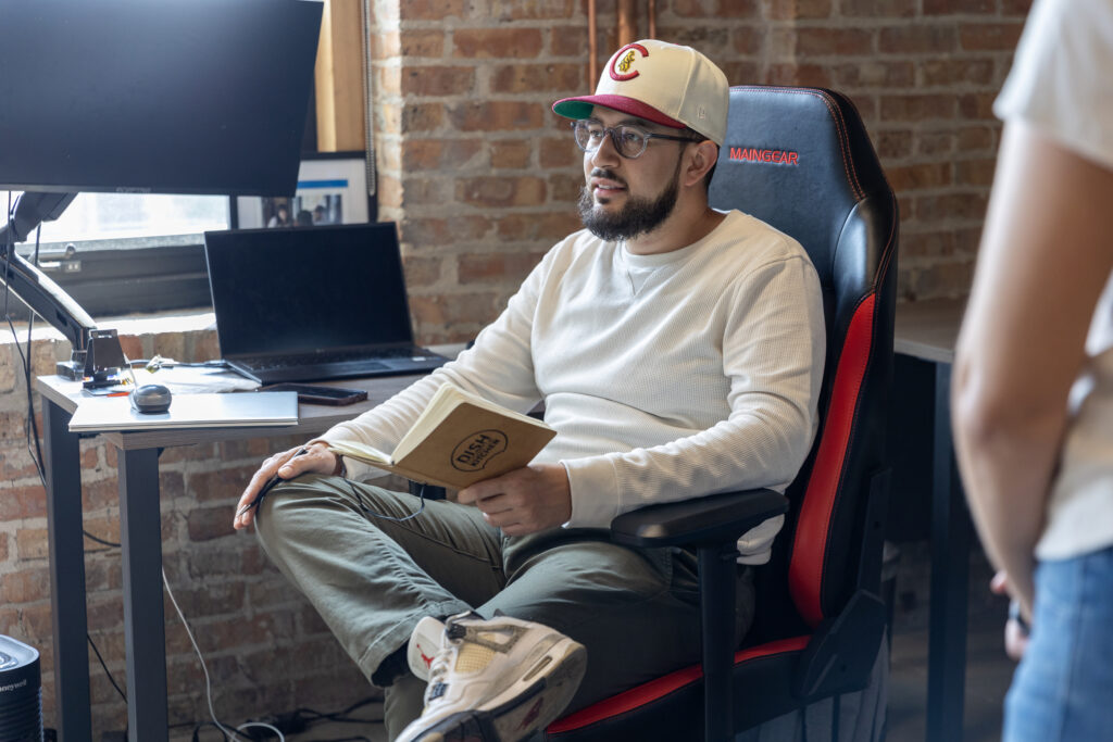 A man wearing a white long-sleeve shirt, a cap, and glasses sits in an office chair holding a notebook with the DishRoulette logo. A desk with a laptop and papers is in the background.