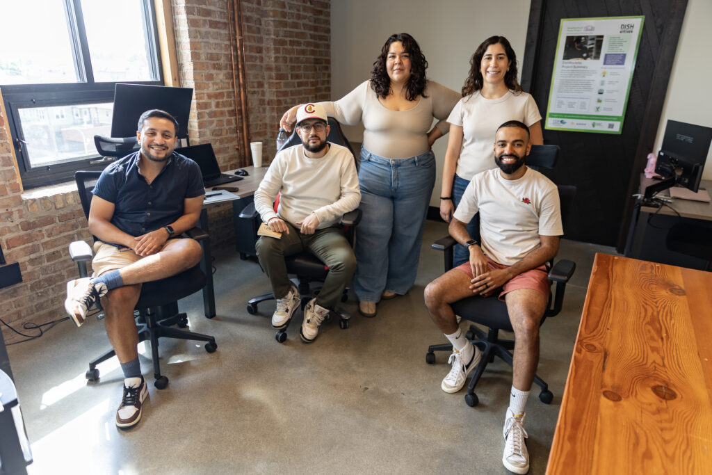 A group of five people smiling and posing in a casual office setting with exposed brick walls, seated and standing near desks and computers.