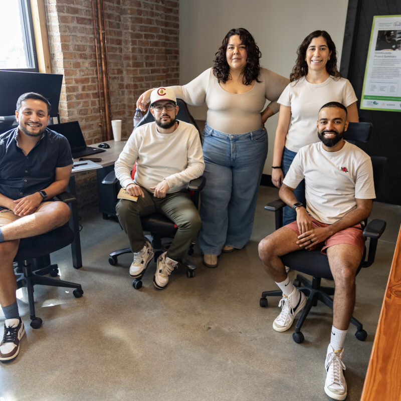 A group of five people smiling and posing in a casual office setting with exposed brick walls, seated and standing near desks and computers.