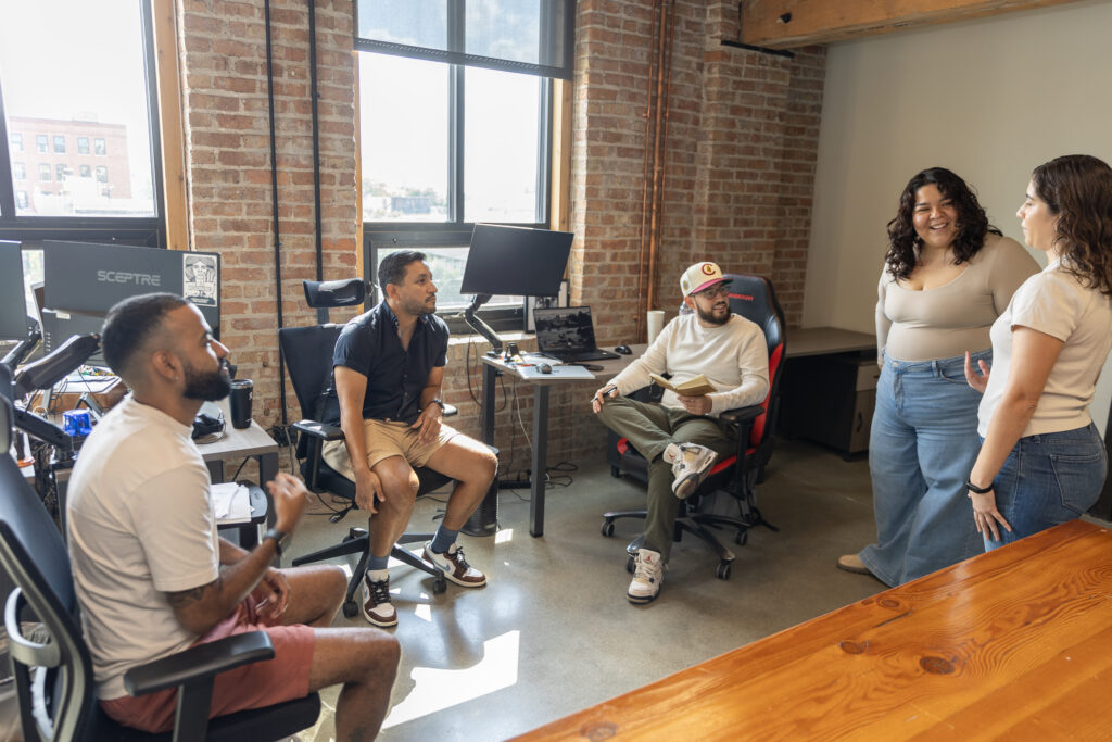 A group of five people in a casual office setting having a conversation, with two standing and three seated near desks and computers in front of an exposed brick wall.