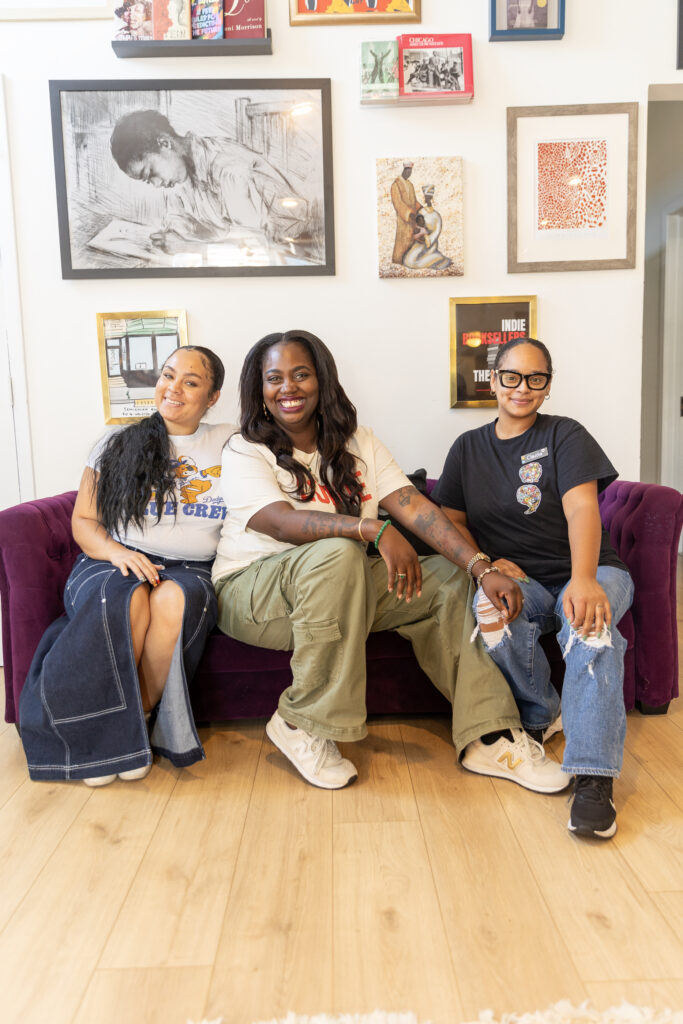 Three women sit on a couch, surrounded by framed artwork on the wall behind them.