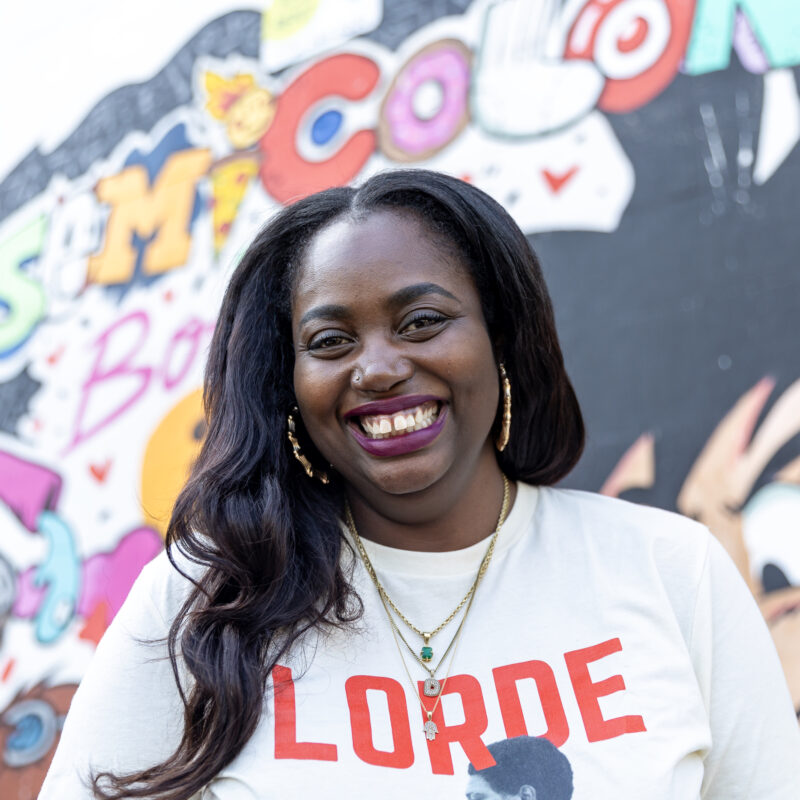 A smiling woman standing in front of a colorful mural at Semicolon Books, wearing a "LORDE" shirt and layered necklaces.