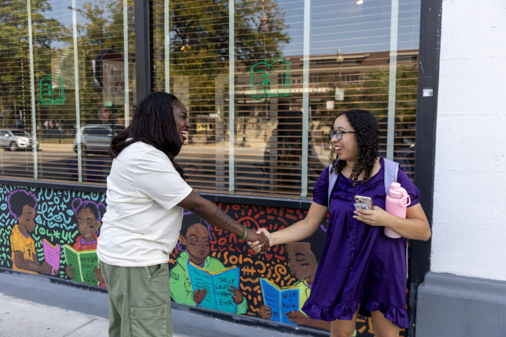 Two women shake hands and smile in front of Semicolon Books, with a colorful mural visible in the background.