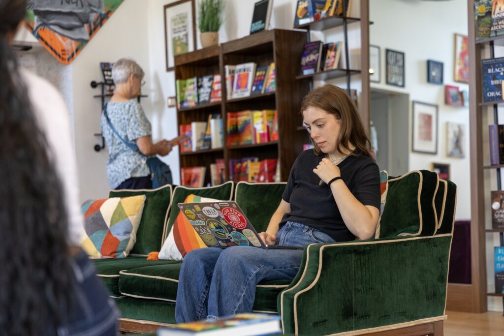 A person sits on a green couch using a laptop while another person browses books in the background.