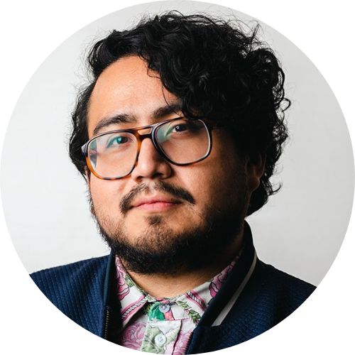 A portrait of a man with curly dark hair and glasses, wearing a patterned shirt and a dark jacket, looking directly at the camera with a neutral expression.