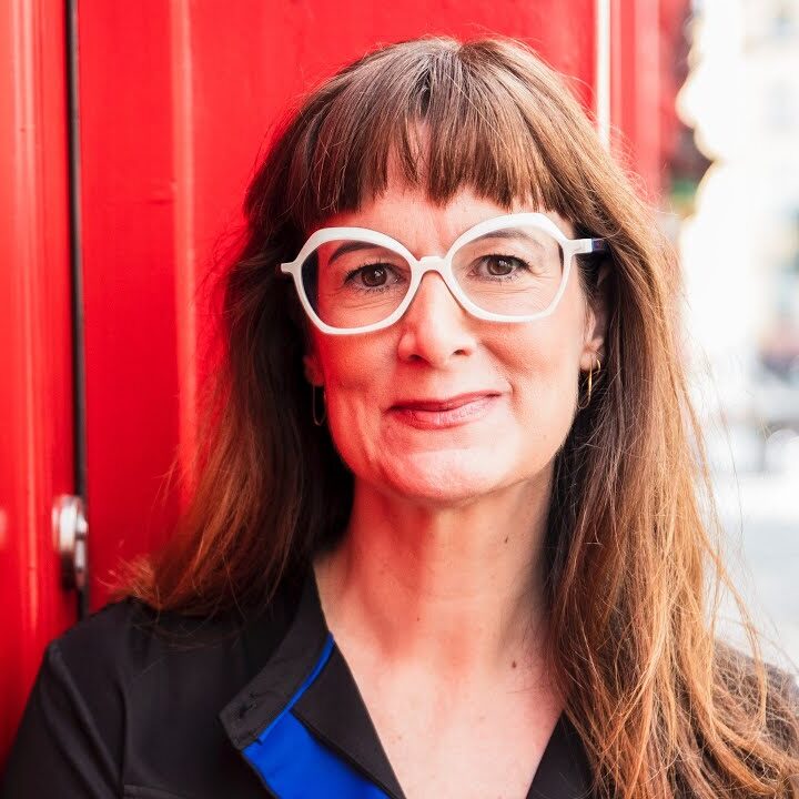 A woman with long brown hair and white-framed glasses stands in front of a bright red wall, smiling