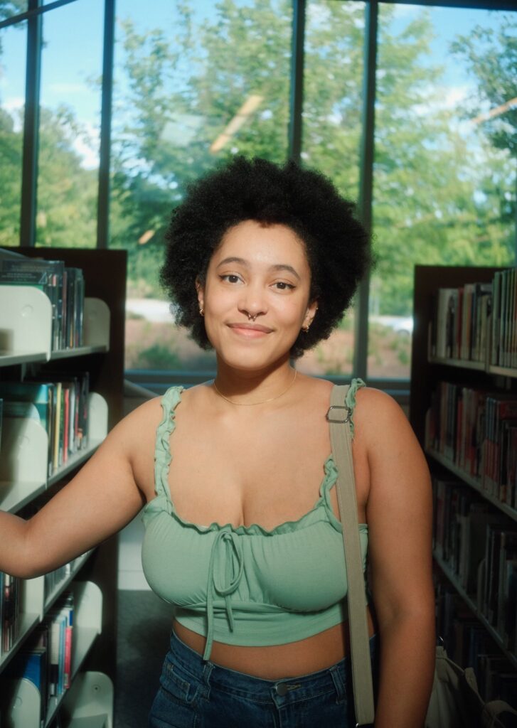 A woman with a wearing a light green top stands confidently between bookshelves at a library.