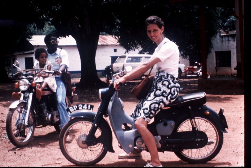 A woman smiles as she sits on a motorbike in the foreground, while in the background, a man rides a motorcycle with a young child sitting in front of him. They are outdoors on a dirt road, with trees and buildings in the background.