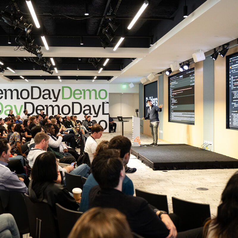 Room filled with people listening to a speaker on stage. Demo Day visual image in the background.