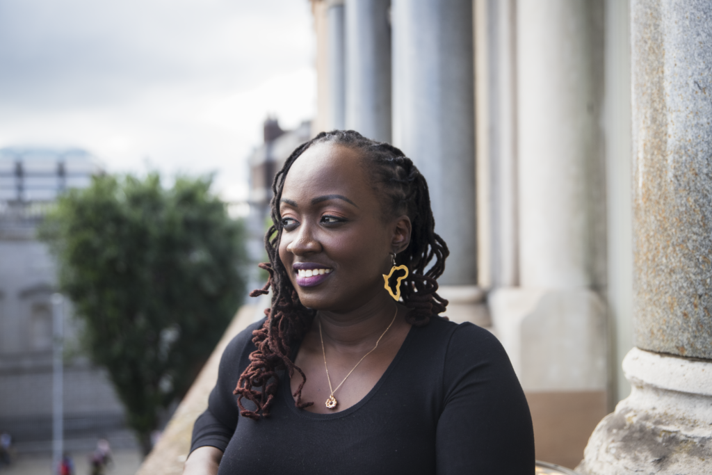 A woman with braided hair and gold Africa-shaped earrings smiles while leaning on a balcony, with columns and greenery in the background.