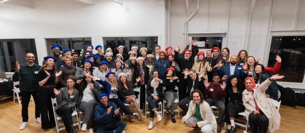 Group photo of attendees at the Columbia Convening on AI Openness and Safety, smiling and waving while wearing blue, red, and white berets, seated and standing in a brightly lit room with large windows.