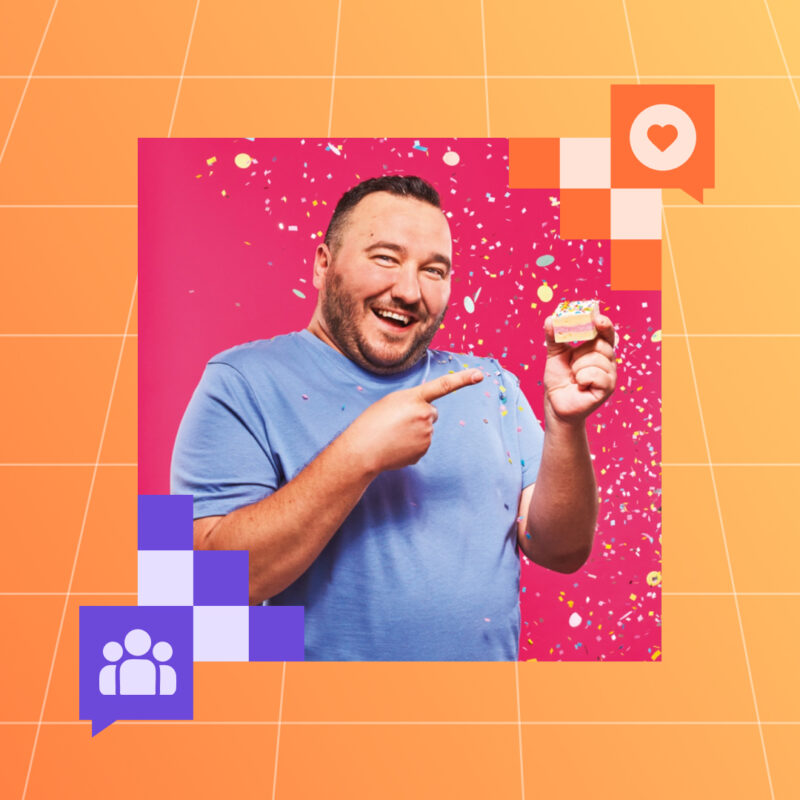 A smiling man in a light blue shirt stands in front of a bright pink background, surrounded by colorful confetti. He’s pointing enthusiastically at a small, square sprinkle-topped cake he’s holding in one hand.