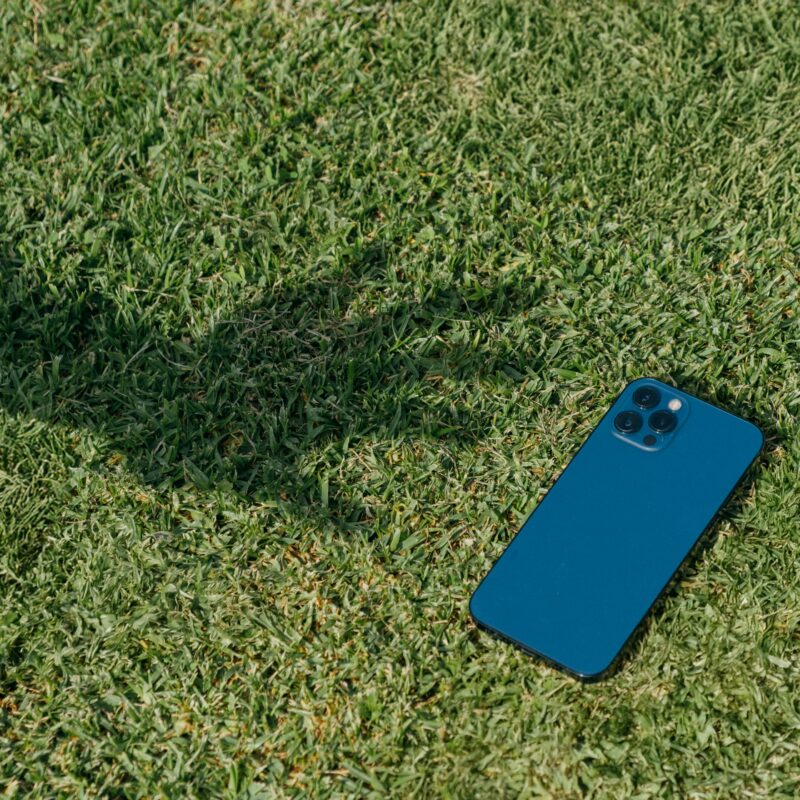 A teal smartphone with triple rear cameras lies on green grass, next to the shadow of an outstretched arm.