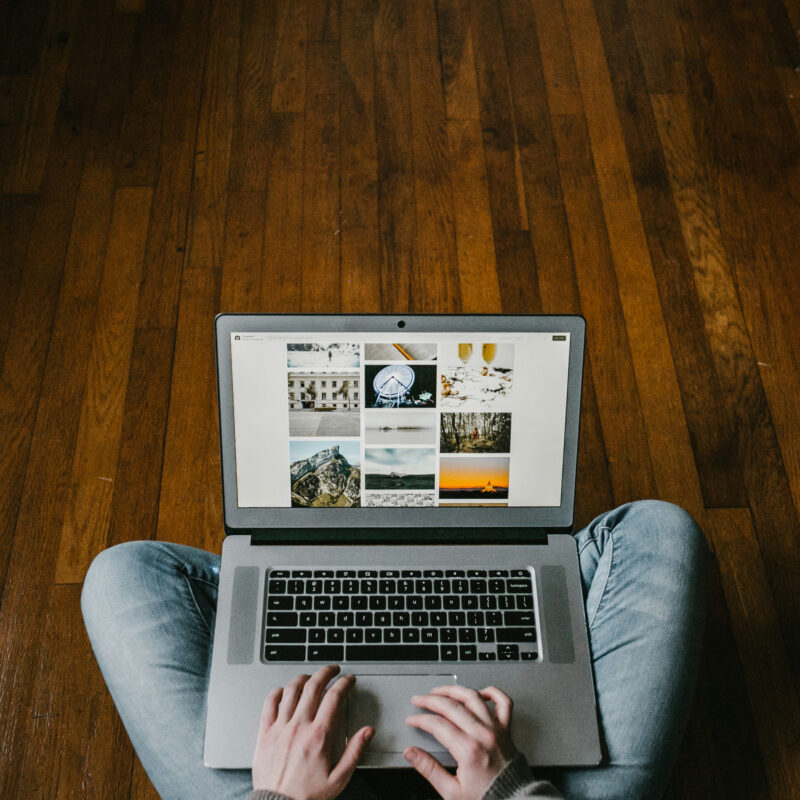 Person browsing photo gallery on laptop while sitting on wooden floor.